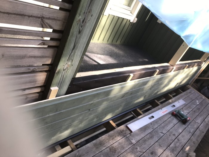 Pågående arbete med liggande träpanel på husvägg och balkongmatta på golv, verktyg och träplank synliga.