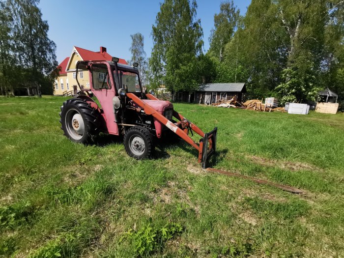 Röd gammal traktor på en lummig gård med gul husfasad och träd i bakgrunden.