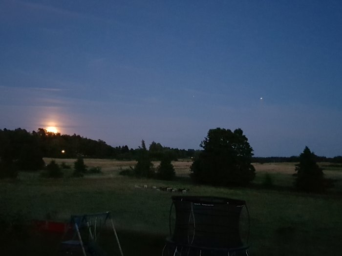 Månen stiger över landskap med träd och fält, ljus på himlen till höger, trädgårdsmöbler i förgrunden.