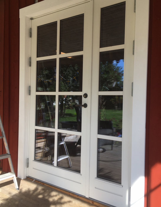 Nyinstallerad vit dörr med glasfönster och dörrfoder på en röd byggnad, och en stege syns reflekterad i glaset.