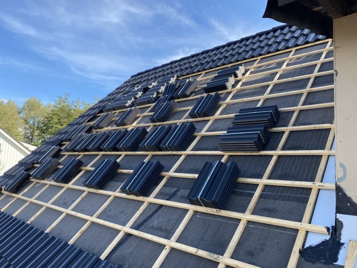 Tak under konstruktion med exponerad takstol och delvis lagda takpannor mot en klarblå himmel.