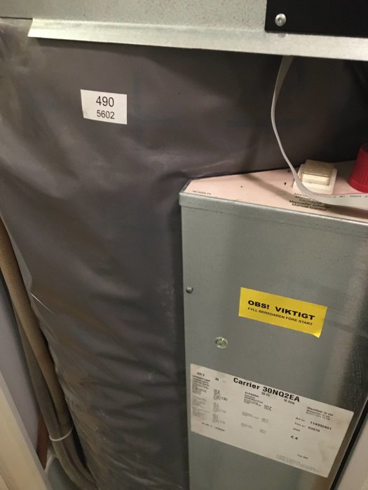 Bild på en Carrier-värmepump med en varningsetikett och specifikationslapp synlig på enhetens sida.