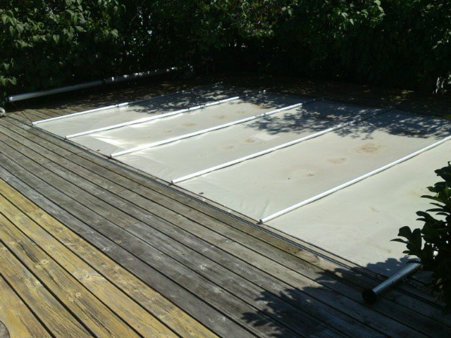 Hemmagjord pooltäckning med beige pressening och aluminiumrör på en trätrall, omgiven av grönska.