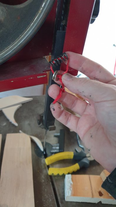 Hand håller en förlorad fjäder framför en bandsåg med verktyg och trästycken i bakgrunden.