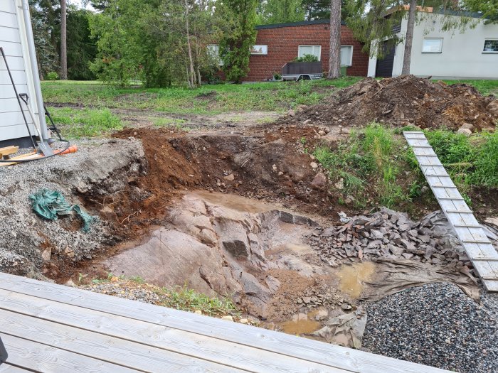 Pågående arbete med grävd poolgrund i trädgård, högar av jord och byggmaterial syns.