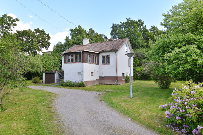 Villa från 1945 med betonghålsten och vitbrun fasad omgiven av en vildvuxen trädgård och grusgång.