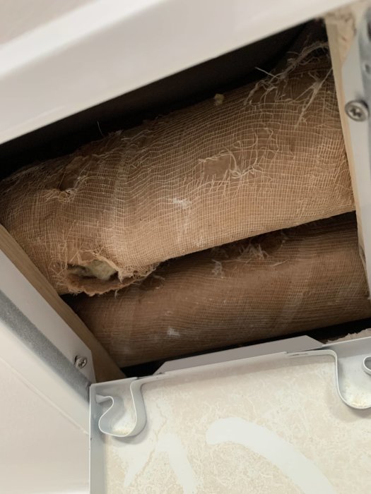 Gammal rörisolering i brunt material syns i slits via inspektionslucka, möjlig asbest, från 1953.