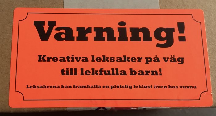 Orange skylt med texten "Varning! Kreativa leksaker på väg till lekfulla barn! Leksakerna kan framkalla en plötslig leklust även hos vuxna".