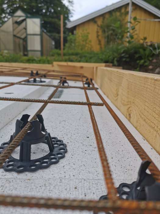 Armeringsnät med distanser på cellplast inför gjutning av betongplatta, med växthus i bakgrunden.