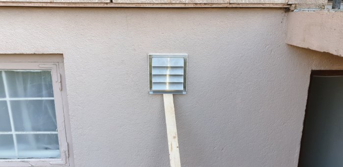 Väggmonterad ventilation med skyddsgaller fastsatt på en träkil vid en husfasad.