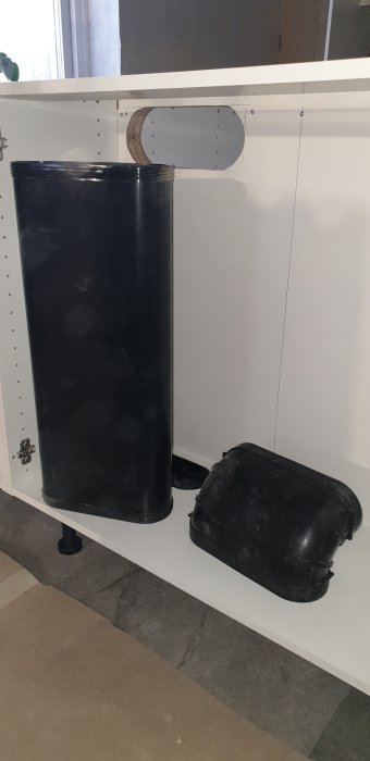 En svart plastbehållare och en svart vattentank placerade under en vit bänk inuti ett rum under konstruktion.