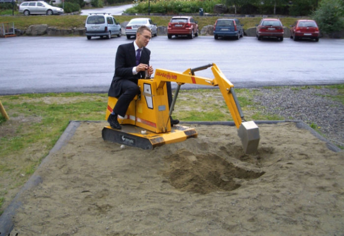 Vuxen man i kostym som leker med en liten grävmaskin i en sandlåda.