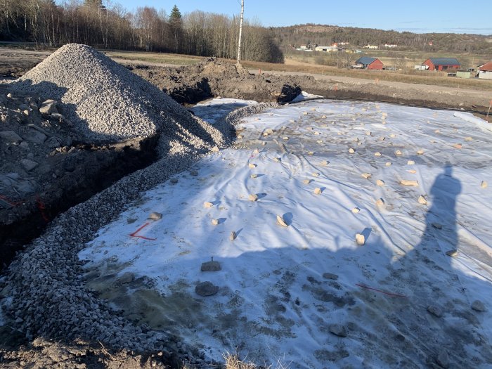 Husgrund med geotextil och bergkross, skugga av en person syns i förgrunden, med landskap i bakgrunden.