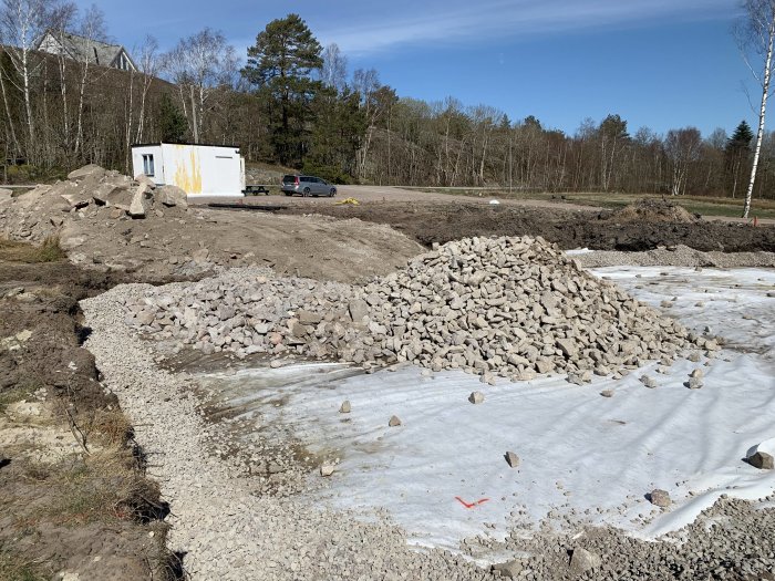 Husgrund och dränering med bergkross och större stenar på byggarbetsplats, byggnadsrelaterade fordon i bakgrunden.