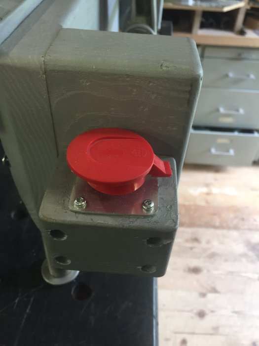 Stor röd brytarknapp monterad på grått maskinstativ med försänkta skruvar.