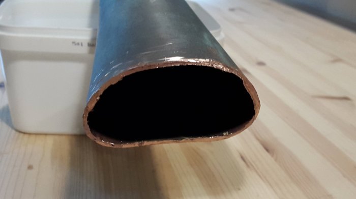 Close-up of a cut and oval-shaped hollow metal pipe end on a wooden surface, part of a repair project.