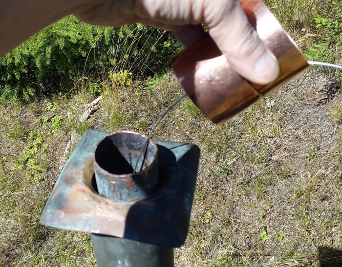 Close-up of a hand holding a copper piece above a metal post with wires, set outdoors in a grassy area, part of a repair process.