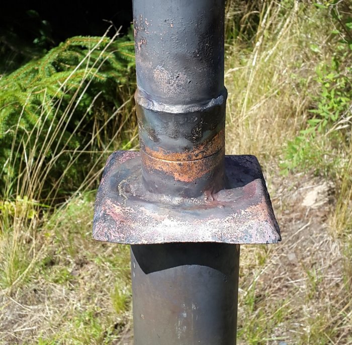 Close-up of a soldered joint on a repaired metal post, with visible welding marks and rust, set outdoors with vegetation in the background.