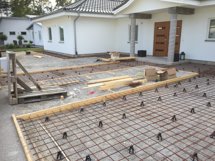 Armeringsnät förberett för betonggjutning framför en villa med byggmaterial omkring.