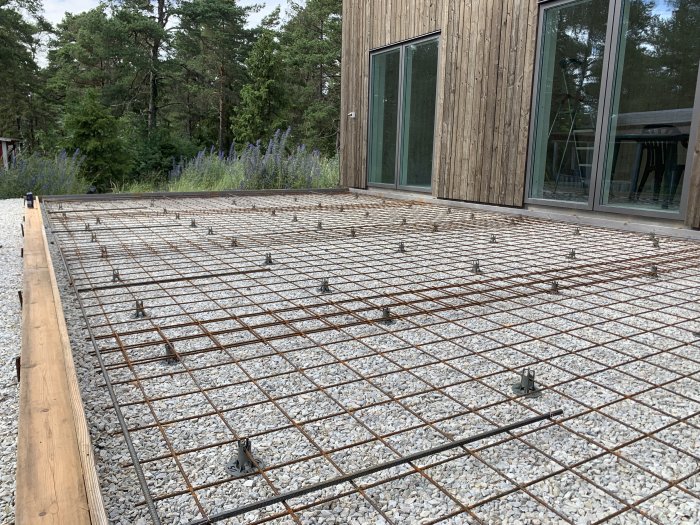 Armeringsjärn och grus i förberedelse för gjutning av betongaltan, med hus och växtlighet i bakgrunden.