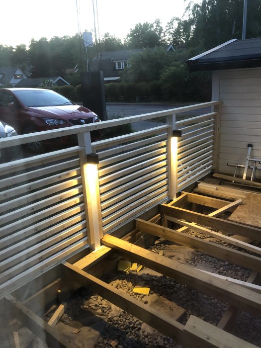 Altan under konstruktion med belysta stolpar och ledningar frästa i träet vid skymning.