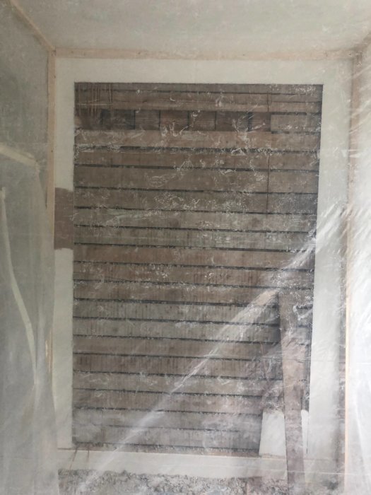 Vägg under renovering där yttre skiktet är avlägsnat, exponerande tegel och reglar, med skyddsplast.