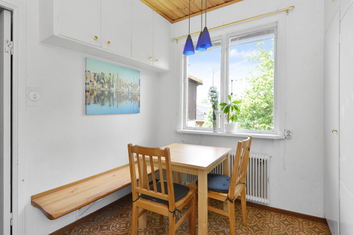 Matplats med träbord och bänk, furutak, en tavla med stadsmotiv, fönster med utsikt och radiator.