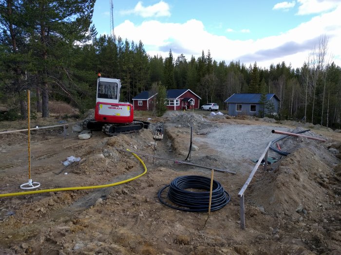 Byggplats för nytt fritidshus med grävmaskin, utgrävd mark och material synligt, befintliga stugor i bakgrunden.