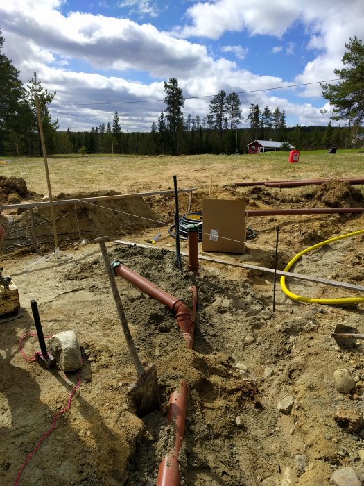 Byggarbetsplats för fritidshus med synligt rörarbete för avlopp och utsikt mot en skog och det gamla röda huset.