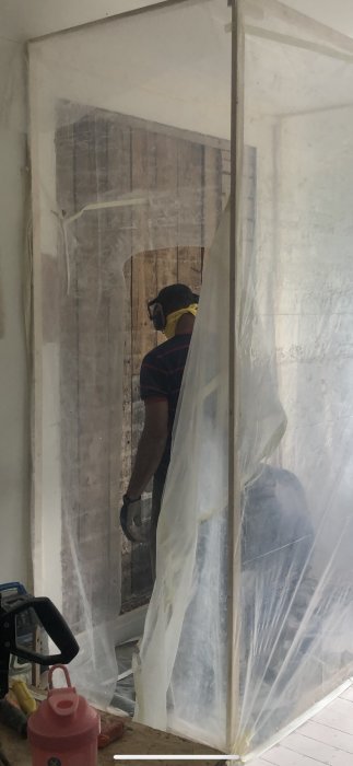 Person med hörselskydd arbetar bakom genomskinlig plast inomhus vid en renovering.
