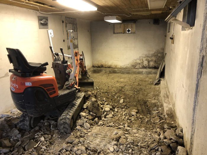 Minigrävare och minidumper i ett garage under byggnation med en uppbilad betongplatta och grushög vid väggen.