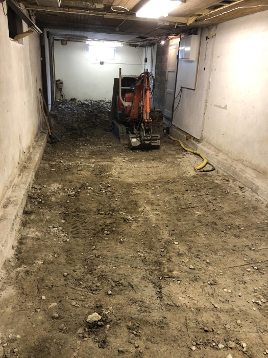 Minigrävare inuti en byggnad under renovering med bortsprängd betongplatta och jordhög.