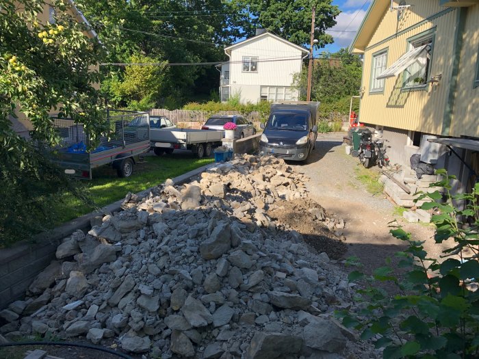 En hög av grus och sten framför ett hus, material från grävningsarbete, med fordon parkerade.