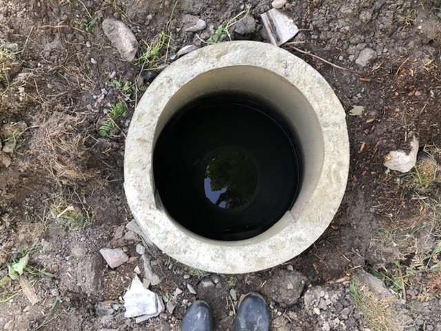Vy ovanifrån av en nyligen höjd betongring i marken, med en spegling av träd i vattenansamlingen inne i ringen.