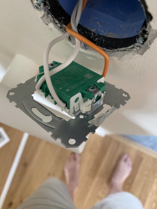 Öppen väggströmbrytare med elledningar synliga, installation i hemmiljö med parkettgolv och en persons fötter.