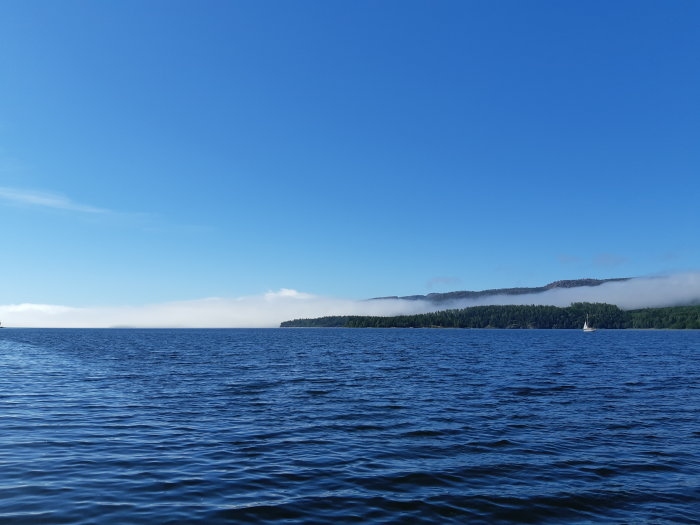 Dimma över skogsbeklädda berg bakom en sjö med segelbåt under en klarblå himmel.