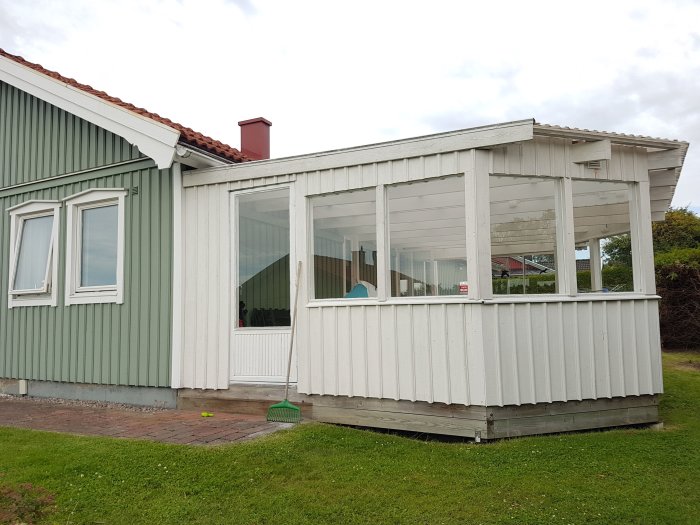 Ett vitt uterum med stora fönster ihopbyggt med ett hus med gröna väggar och vitt tak.