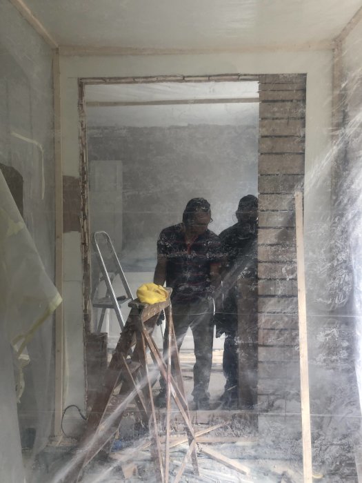 Två personer i en dammig byggarbetsplats med plasttäcke och en stege i förgrunden.