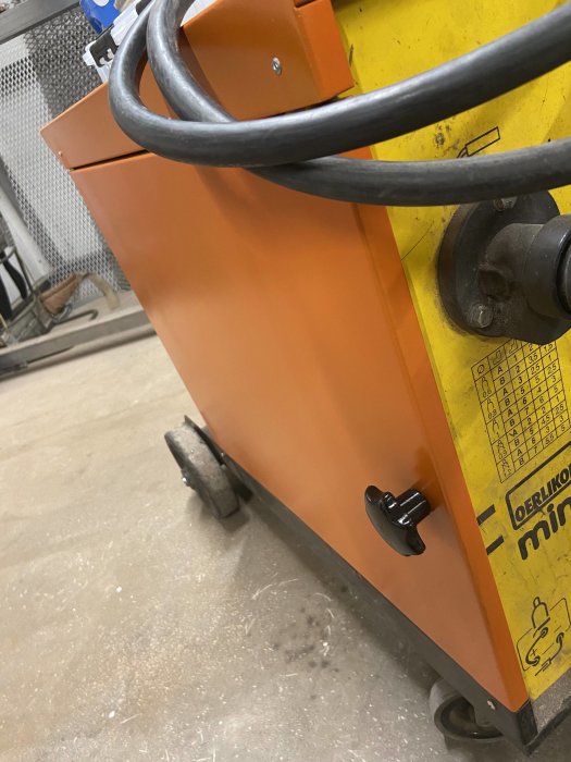 Nymålad orange och gul svetsmaskin med ny slang och låsratt, står på betonggolv.