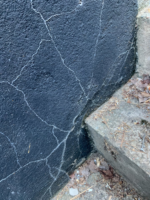 Yttervägg med spruckna och lossnande delar av svart puts vid en källarnedgång.