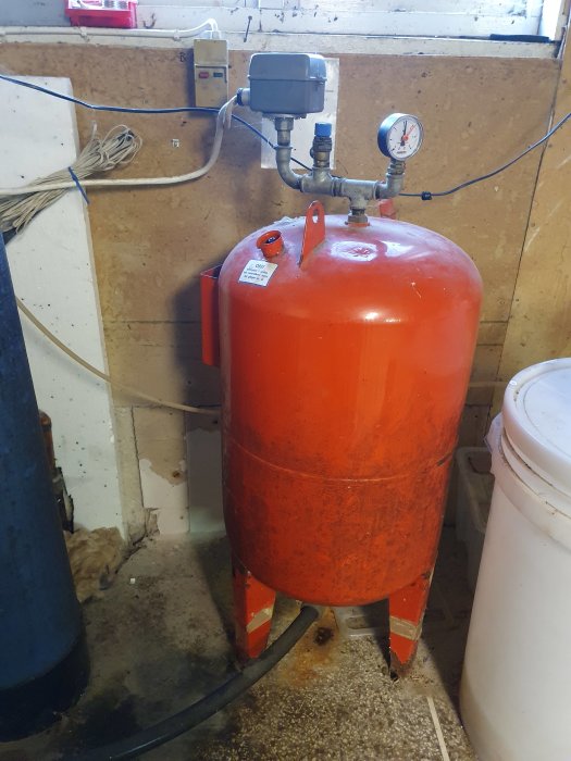 Hydropress i rött med manometer på toppen och en slang från botten, placerad i ett verkstadsrum.