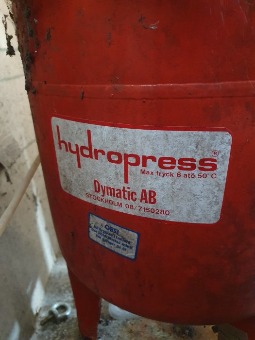 En röd hydropress från Dymatic AB med varningsetikett, som är ungefär 90cm hög, med slitage och utan synlig manometeranslutning.