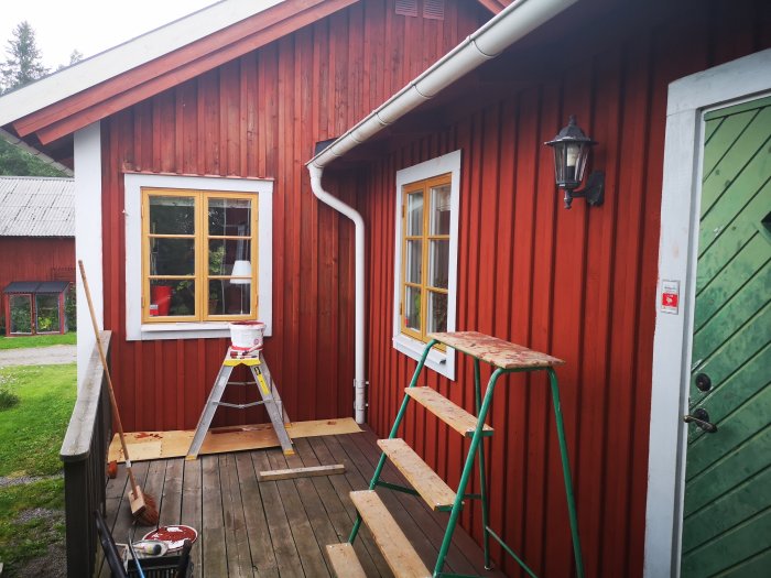 Rödmålat trähus med stegar och målarutrustning på verandan, färska målningsarbeten synliga.