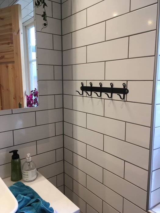 Handdukskrokar på vit kaklad badrumsvägg över en handduk och tvålflaskor.