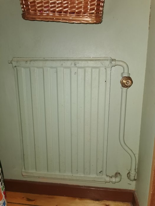 Äldre vit radiator utan vred med synlig liten pinne, rörkopplingar och avtappningsventil i ett hus.