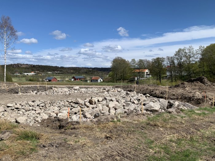 Ett område med stora utlagda stenar redo för vältning vid ett byggprojekt, med hus och träd i bakgrunden.