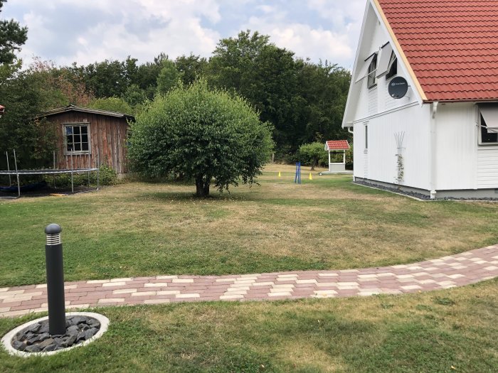 Vissnad gräsmatta nära ett vitt hus, brun lekstuga och gröna buskar under sommarvärme.