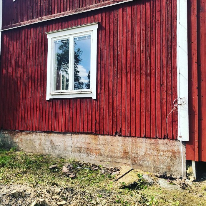 En rödmålad timmerbyggnad med grunden delvis grävd framför och synlig putsad yta undertill.