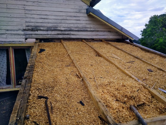 En avtäckt husgrund täckt med spån och några spadar i förgrunden, med ett hus och träd i bakgrunden.