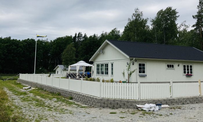 Ny målad staket framför hus med svensk flagga, oavslutade stolpar och byggmaterial på marken.